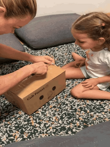A woman and a young girl sit on a patterned carpet, opening a cardboard box printed with the brand name 'Cub Forest'.